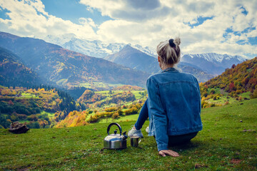 Young woman in jeans with a teapot and a cup of hot coffee is sitting on the edge and watching an amazing beautiful mountain landscape on an autumn day. Concept of travel, camping, outdoor activities