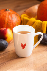 white cup with a red heart pattern of tea with an autumn decor of pumpkins, plums, textiles and peaches on a wooden background