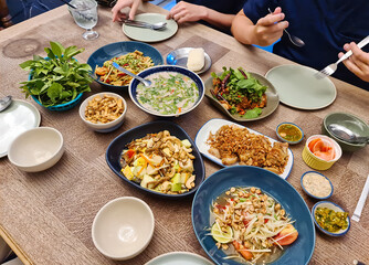 Variety of Thai food with papaya spicy salad, fruit salad, crispy pork, spicy pork soup, sticky rice and cutlery on wooden table