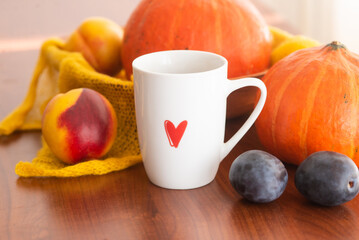 white cup with a red heart pattern of tea with an autumn decor of pumpkins, plums, textiles and peaches on a wooden background