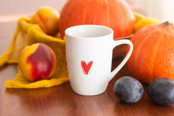 white cup with a red heart pattern of tea with an autumn decor of pumpkins, plums, textiles and peaches on a wooden background