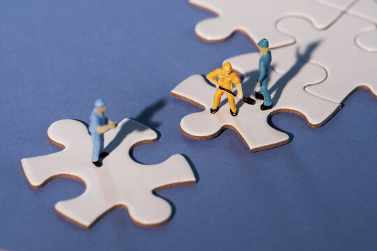 Team of tiny worker miniature figures on linked jigsaw puzzle pieces island on dark blue, purple paper. Close-up on white puzzle elements linked together and separate. Dramatic light, long shadows.