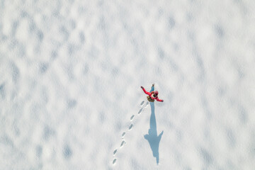 Woman in a red christmas jacket with backpack walking on the snow and doing footprints in a park. Winter. Drone, top, aerial view.