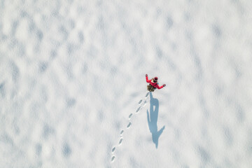Woman in a red christmas jacket with backpack walking on the snow and doing footprints in a park. Winter. Drone, top, aerial view.