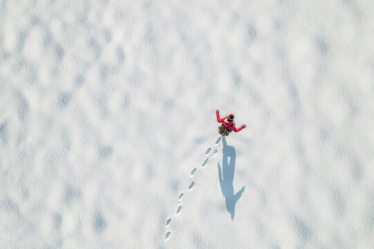 Woman in a red christmas jacket with backpack walking on the snow and doing footprints in a park. Winter. Drone, top, aerial view.