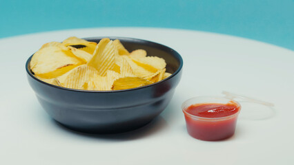 crunchy and ridged potato chips in bowl near plastic container with ketchup on blue.
