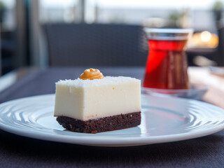 slices of snow white milk agar gelatin dessert (pudding, mousse, souffle) with coconut flakes on chocolate cake islak kek on white plate and turkish tea in tulip shape glass in outdoor cafe