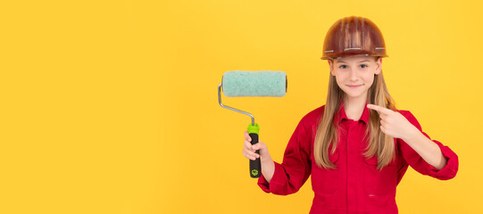 happy teen child in builder hard hat with paint roller on yellow wall. pointing finger. Child builder in helmet horizontal poster design. Banner header, copy space.