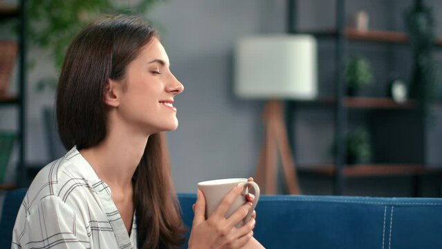 Happy female drinking hot tea coffee beverage dreaming enjoy break relaxing on blue couch at home