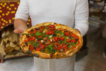 Pizza napoletana con pomodorini, basilico fresco, aglio, origano e olio sorretta da un pizzaiolo napoletano davanti al forno al legna di una pizzeria