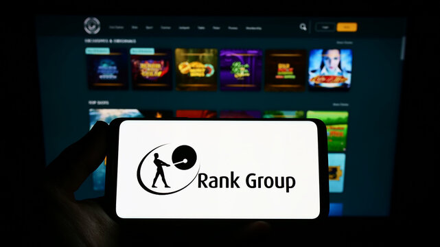 Stuttgart, Germany - 09-15-2022: Person holding smartphone with logo of British gambling company The Rank Group plc on screen in front of website. Focus on phone display.