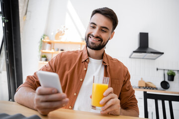 cheerful and bearded man holding glass of orange juice while using smartphone.