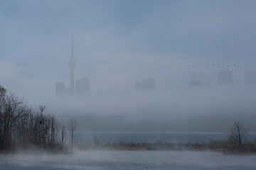 Fototapeta premium Fog and mist over lake Ontario water surface covers Toronto city skyline, heavy clouds on the sky. Weather conditions, advisory, alert concept. Space for copy.