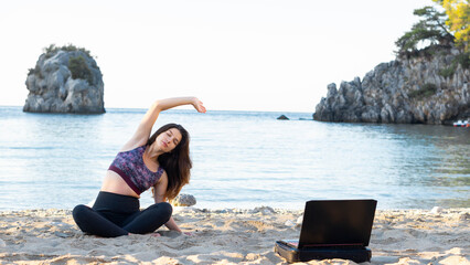 Fitness and yoga instructor holding online training via laptop. Showing yoga exercises at the beach in front of  laptop,  Yoga and meditation concept. Virtually hobby time concept.