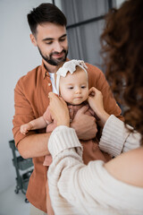 curly mother adjusting headband of baby daughter near cheerful husband at home.