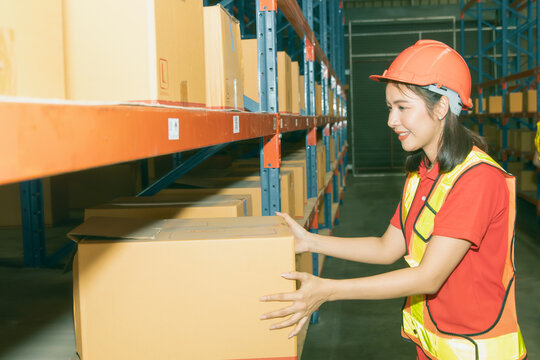 Side View Beautiful Young Asian Female Worker Working The Night Shift Wearing A Helmet Works With Smiling Face, Use Tablet To Inspect The Boxes From The Warehouse Shelf To Verify They Are Correct.