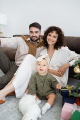 happy parents and cheerful infant girl sitting near decorated christmas tree with presents.