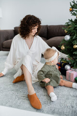 curly mother sitting near decorated christmas tree with presents and infant daughter.