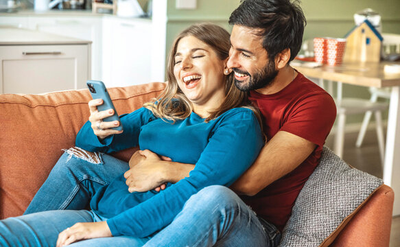 Happy Couple Watching Smart Mobile Phone Device Sitting On Sofa At Home - Husband And Wife Having Fun Together Using Cellphone - Happy Lifestyle And Technology Concept