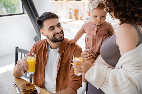 curly woman holding in arms infant baby near smiling husband with glass of orange juice.