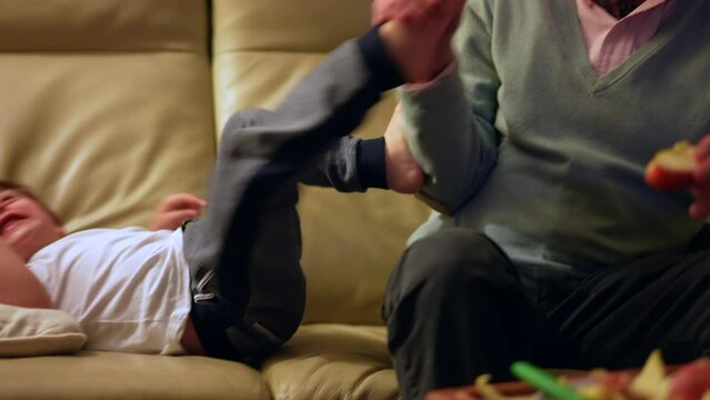 Child misbehaving hitting grandfather with feet. Disobedient little boy misbehaves on couch with grandparent