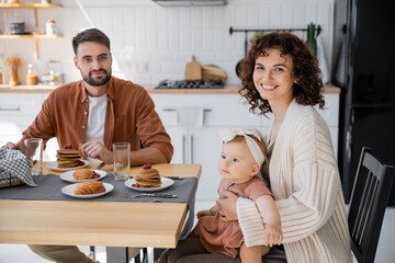 cheerful mother holding infant daughter and sitting at table with husband during breakfast.