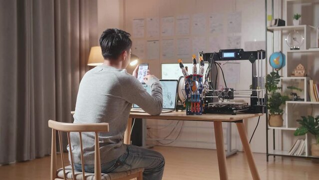 Back View Of A Male With 3D Printing Looking At The Pictures On Smartphone While Designing A Cyborg Hand On A Laptop At Home
