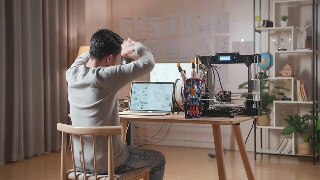 Back View Of A Male With 3D Printing Stretching  While Designing A Cyborg Hand On A Laptop At Home
