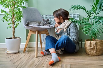 Cold winter season, woman with cat on chair near heating radiator