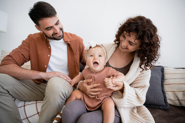 cheerful mother holding amazed infant daughter near bearded husband sitting on couch.
