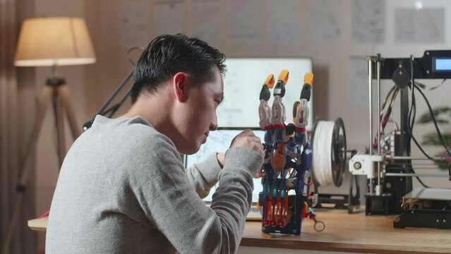 Side View Of Asian Male With 3D Printing Repairing A Cyborg Hand While Designing It On A Laptop At Home
