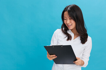 Fototapeta premium Happy cute Asian businesswoman in classic office dress code holds folder tablet read candidate's resume posing isolated on over blue studio background. Cool business offer. Job interview concept
