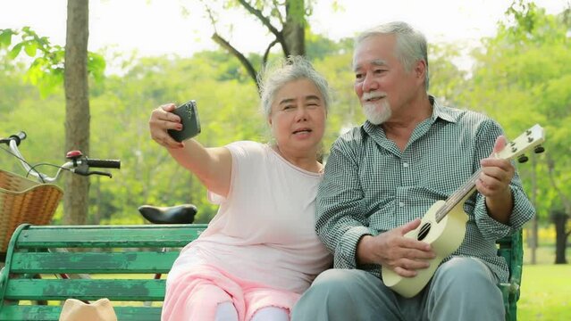 Elderly asian couple sitting do recreational activities together in the garden, spending their free time taking selfies, singing and having fun playing the ukulele.