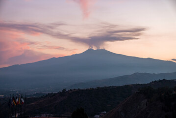 Fototapeta premium Smoke emitting from Mount Etna with orange sky in background. Beautiful tourist attraction with luxurious Elios hotel in foreground. Scenic view of dramatic landscape during sunset.