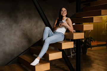Young asian woman in eyeglasses using cellphone while sitting on stairs