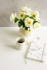 Feminine lifestyle composition, minimal workspace neutral colors. Vase with beige roses flowers , cup coffee,diary,pen on white desk near  gray background with copy space .Breakfast stationary mockup.