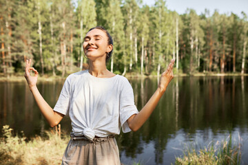 Portrait of young woman practicing yoga outdoor standing on background with river bank of coniferous forest, breathing deeply inhaling pure fresh air, doing relaxing, stress relief techniques