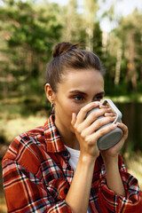 Woman in red checkered shirt drink coffee from white mug in wild nature standing outdoor with lake and forest on background, enjoying tranquility and peace, fresh air of summer woodland