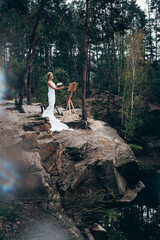 Fototapeta premium Art work. A slender woman with a stylish haircut, wrapped in a white long material, stands with a brush in her hand, on the edge of a picturesque cliff and draws with oil paints on an easel.