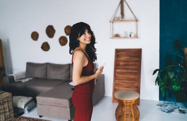 Young woman with smartphone standing near sofa in room