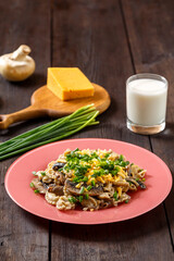 Pasta in a creamy sauce with mushrooms in a plate on a dark table next to a glass of cream, cheese on a wooden board, green onions and champignons.