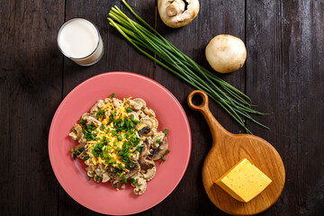 Pasta in a creamy sauce with mushrooms in a plate on a dark wooden table next to a glass of cream, cheese on the board, green onions and mushrooms top view.