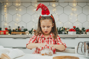 Little dark-haired girl 3 years old in red Christmas cap and checkered shirt prepares dough for gingerbread cookies in white Christmas decorated kitchen with garland lights. Merry Christmas, New Year