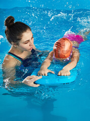 Little girl learning to swim in indoor pool with pool board and trainer