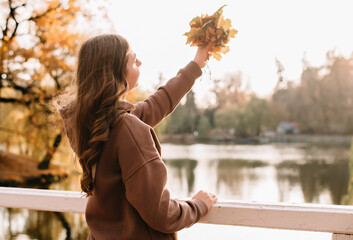 Young woman in hoodie holding autumn leaves a in an autumn park. Sunny weather. fall season.