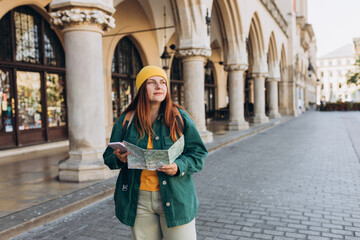 Attractive young female tourist is exploring new city. Redhead girl in hat holding a paper map and smartphone on Market Square in Krakow. Traveling Europe in autumn. Active lifestyle concept