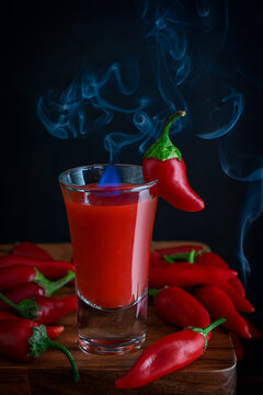 Single Portion Of Red Alcohol Hot Cocktail Shot Drink Made Of Tomato Juice, Chili Pepper Sauce And Vodka Or Tequila Served In Glass With Blue Flame And Smoke On Dark Brown Wooden Bar Counter At Club