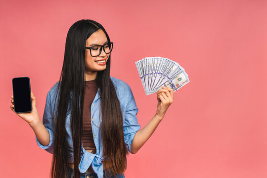 Happy Rich Winner! Portrait Of Asian Woman Holding Money Dollars Bills And Smiling Satisfied, Standing Isolated Over Pink Background. Using Mobile Phone. Showing Screen.