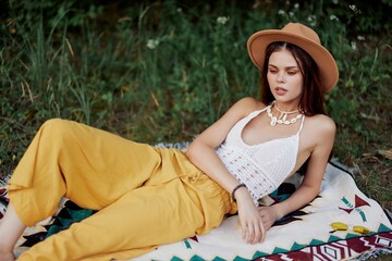 A woman in eco-friendly clothing in a hippie look is lying on a colored plaid smiling and looking at an autumn sunset in nature in the park. Lifestyle on a journey of harmony with the world