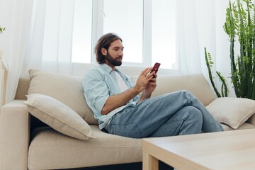 Obraz premium A man with a beard sits on the couch during the day and smiles at home and looks at his phone relaxing on his day off, life online on his phone texting with friends